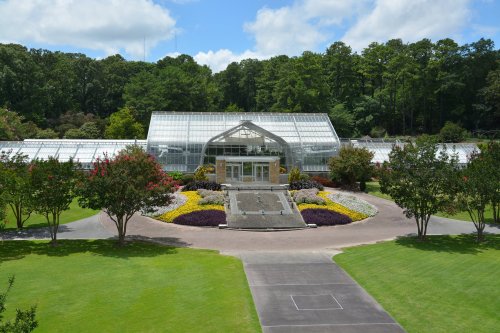 A large glass greenhouse surrounded by colorful flower beds and lush greenery under a blue sky.