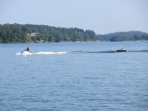 A person on a jet ski tows another person on a tube across a calm lake surrounded by trees.