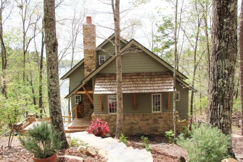 A charming green cottage with a stone chimney, surrounded by trees and colorful flowers in a serene outdoor setting.