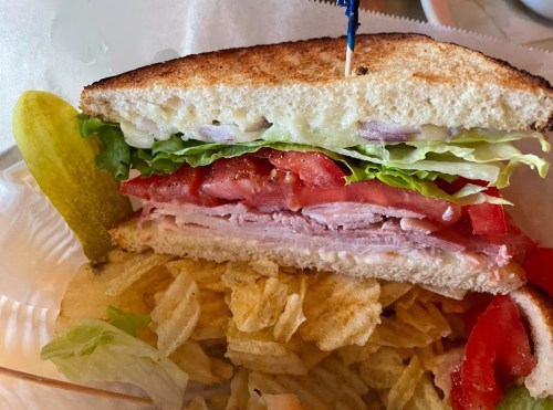 A close-up of a sandwich with layers of turkey, lettuce, tomato, and onions, served with a pickle and potato chips.