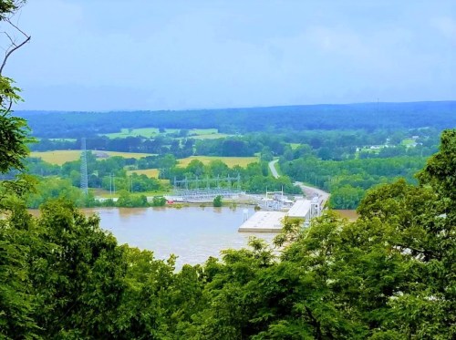 A scenic view of a river and dam surrounded by lush greenery and rolling hills under a cloudy sky.