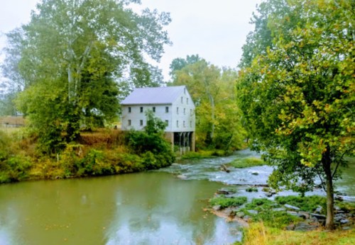 A historic white mill stands by a calm river, surrounded by lush green trees and a serene landscape.