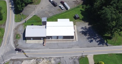 Aerial view of a large building with a white roof, surrounded by a parking lot and greenery along a road.