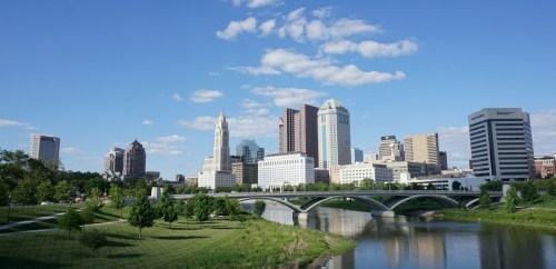 A skyline view of Columbus, Ohio, featuring tall buildings and a river under a clear blue sky.