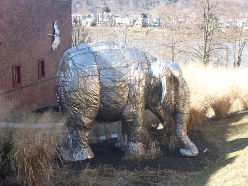 A large metallic elephant sculpture stands among tall grass near a brick building, with a bird flying above.