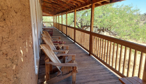 A wooden porch with several chairs, overlooking a natural landscape with trees and dry ground.