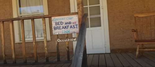 Sign on a porch reads "Kentucky Camp Bed and No Breakfast Inn" with "Occupied" below it. Wooden railing in foreground.