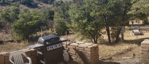 A barbecue grill next to a stone wall, surrounded by trees and a hilly landscape.
