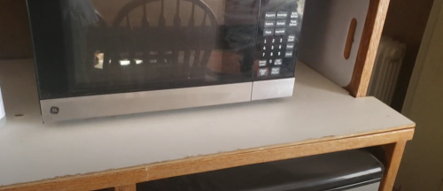 A microwave on a wooden shelf, with a chair and a black appliance visible in the background.