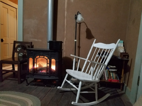 A cozy room with a white rocking chair, a wood stove, and a lamp, creating a warm and inviting atmosphere.