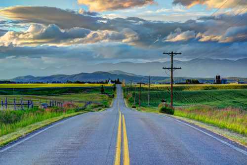 A winding road stretches into the distance, flanked by fields and mountains under a cloudy sky.