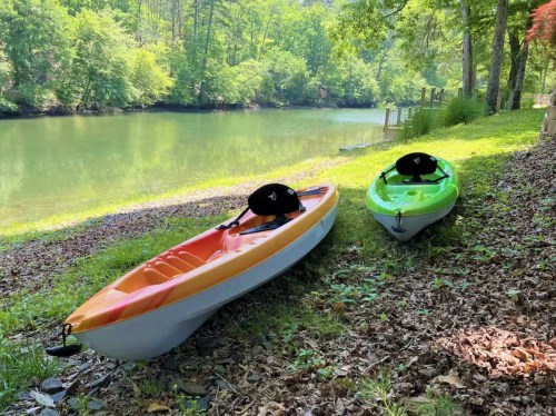 Two kayaks, one orange and one green, sit on the grassy bank by a calm river surrounded by trees.
