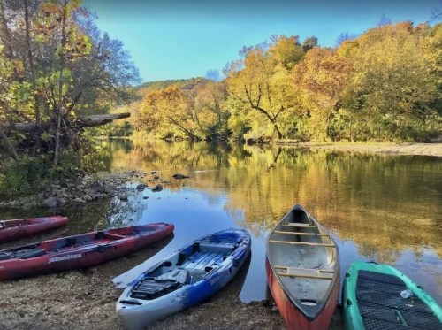 Colorful kayaks and canoes are moored by a serene river surrounded by autumn foliage and clear blue skies.