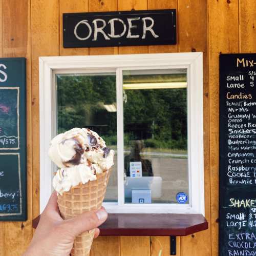 A hand holds an ice cream cone in front of a wooden stand with a sign that says "ORDER" and a menu displayed.