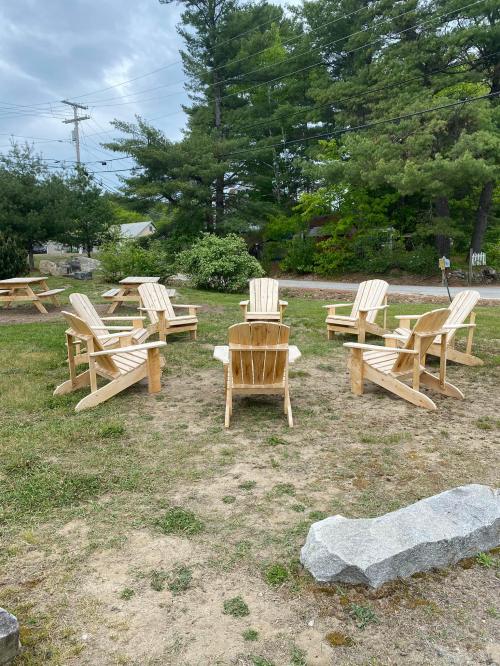 A circle of wooden Adirondack chairs on a grassy area, surrounded by trees and a pathway.