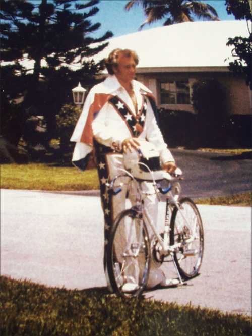 A person in a star-patterned jumpsuit stands next to a bicycle in a suburban setting with palm trees.