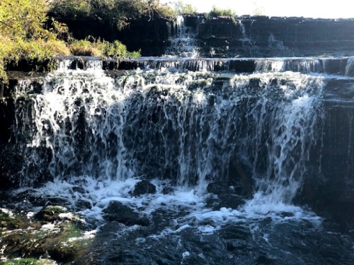 A cascading waterfall flows over rocky terrain, surrounded by lush greenery and sunlight.