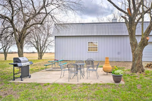 A patio with a grill, table, and chairs beside a gray building and trees, set in an open field.