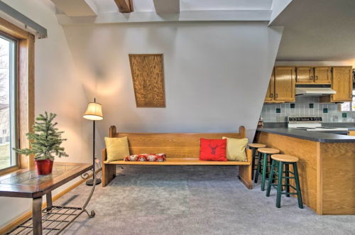 Cozy living space with a wooden bench, decorative pillows, a small tree, and a kitchen area in the background.