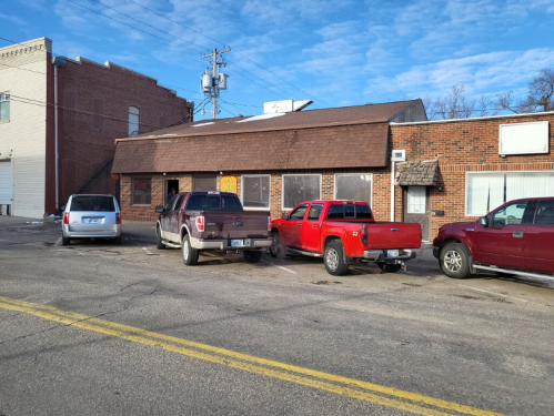 A small brick building with a brown roof, featuring parked vehicles in front and a clear blue sky above.