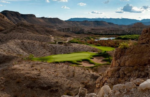 A scenic view of a golf course nestled among rocky hills and a river, under a partly cloudy sky.