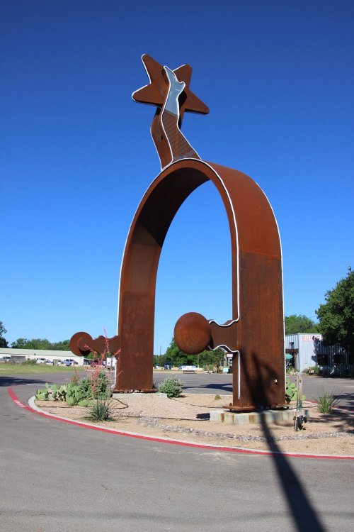 A large, rusted metal sculpture of a star and arch, set against a clear blue sky, surrounded by desert plants.