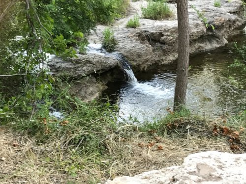 A small waterfall flows over rocks into a calm pool, surrounded by greenery and trees.