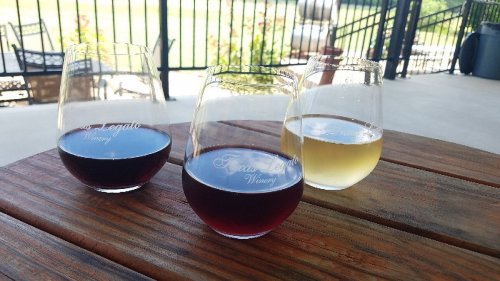 Three wine glasses on a wooden table: two with red wine and one with white wine, set against a sunny outdoor backdrop.