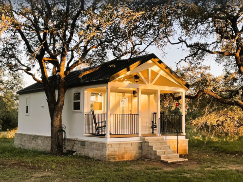 A small white house with a porch, surrounded by trees, illuminated by warm sunlight in a serene outdoor setting.