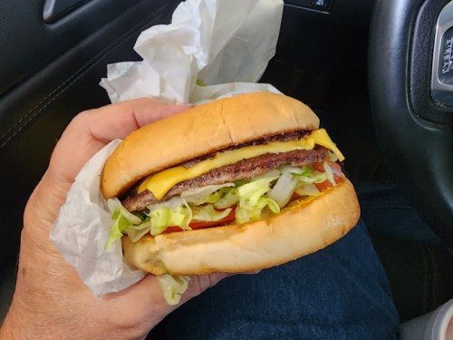 A hand holding a cheeseburger with lettuce, tomato, and cheese, inside a car.