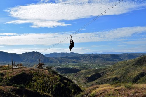A person zip-lining over a scenic landscape of mountains and valleys under a blue sky with scattered clouds.