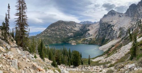 A panoramic view of a serene lake surrounded by mountains and pine trees under a cloudy sky.