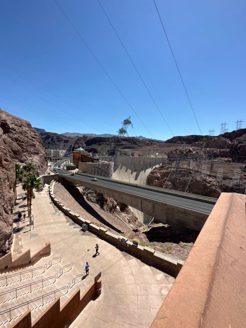 A view of a dam surrounded by rocky terrain and power lines under a clear blue sky, with people walking below.