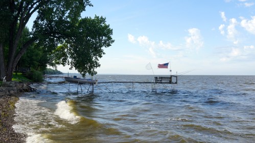 A peaceful lakeside scene with a dock, a boat, and an American flag waving under a blue sky with scattered clouds.