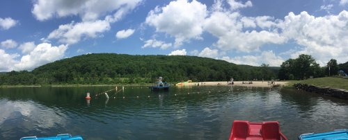 A serene lake scene with a sandy beach, green hills, and fluffy clouds under a bright blue sky.