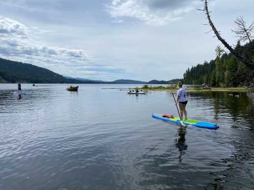 A person paddleboards on a calm lake surrounded by trees and mountains under a cloudy sky.