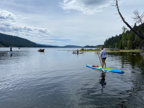A person paddleboards on a calm lake surrounded by trees and mountains under a cloudy sky.