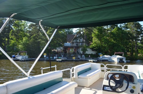 View from a boat showing a lakeside restaurant and dock with boats, surrounded by trees.