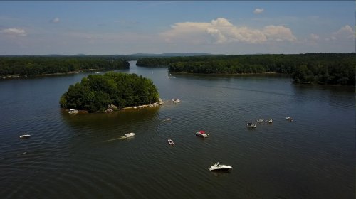 Aerial view of a serene lake with boats and a small island surrounded by lush greenery under a clear blue sky.