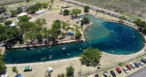 Aerial view of a winding river with people swimming, surrounded by trees and grassy areas, with parked cars nearby.