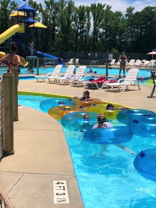 A sunny pool scene with people floating in colorful tubes, a water slide in the background, and lounge chairs nearby.