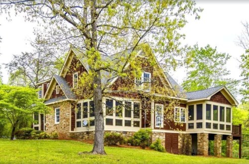 A charming two-story house with a stone foundation, large windows, and a screened porch, surrounded by lush greenery.