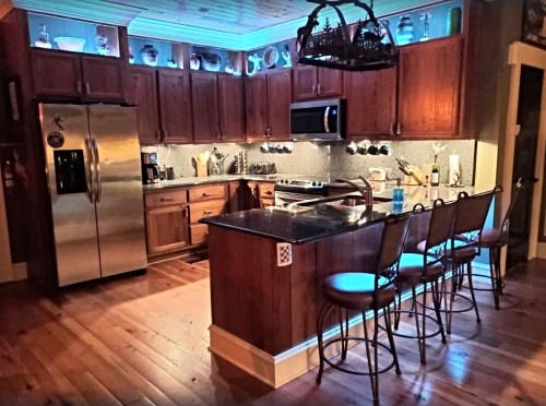 A modern kitchen with wooden cabinets, stainless steel appliances, and a central island with bar stools. Soft blue lighting.