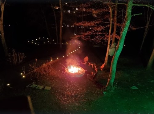 A cozy outdoor scene at night with a fire pit, surrounded by chairs and glowing lights in the trees.