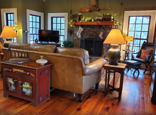 Cozy living room with a stone fireplace, leather sofa, lamps, and festive decorations. Warm wooden floors and windows.
