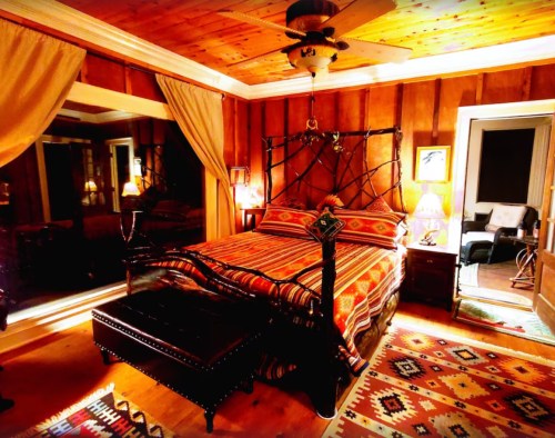 Cozy bedroom with wooden walls, a patterned bedspread, and warm lighting, featuring a ceiling fan and decorative rugs.