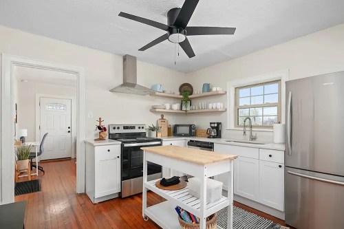 Bright kitchen with white cabinets, stainless steel appliances, a wooden island, and a ceiling fan. Natural light from a window.