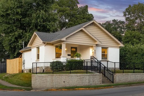 A charming white house with a gray roof, surrounded by greenery and a black fence, at dusk.