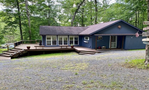 A blue house with a wooden deck surrounded by trees, featuring a gravel driveway and potted plants on the porch.