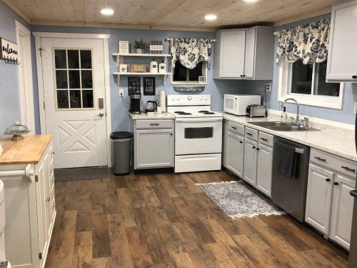 A cozy kitchen with blue walls, white cabinets, and wooden flooring, featuring a stove, sink, and small dining area.